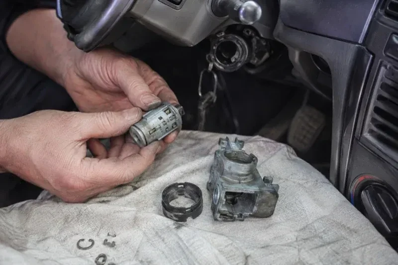 Hands holding a removed ignition cylinder beside a car steering column, with the housing and small parts on a cloth. — Ignition Lock Repair, Toronto